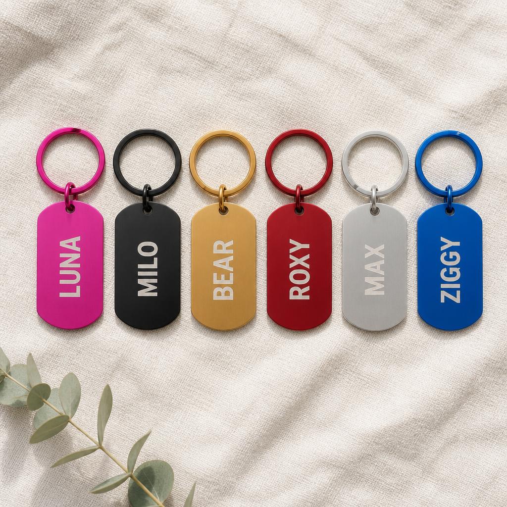 Six personalized engraved keychains in a row — hot pink, black, gold, red, silver, and blue — each with a name engraved as sanded bare aluminum — on cream linen